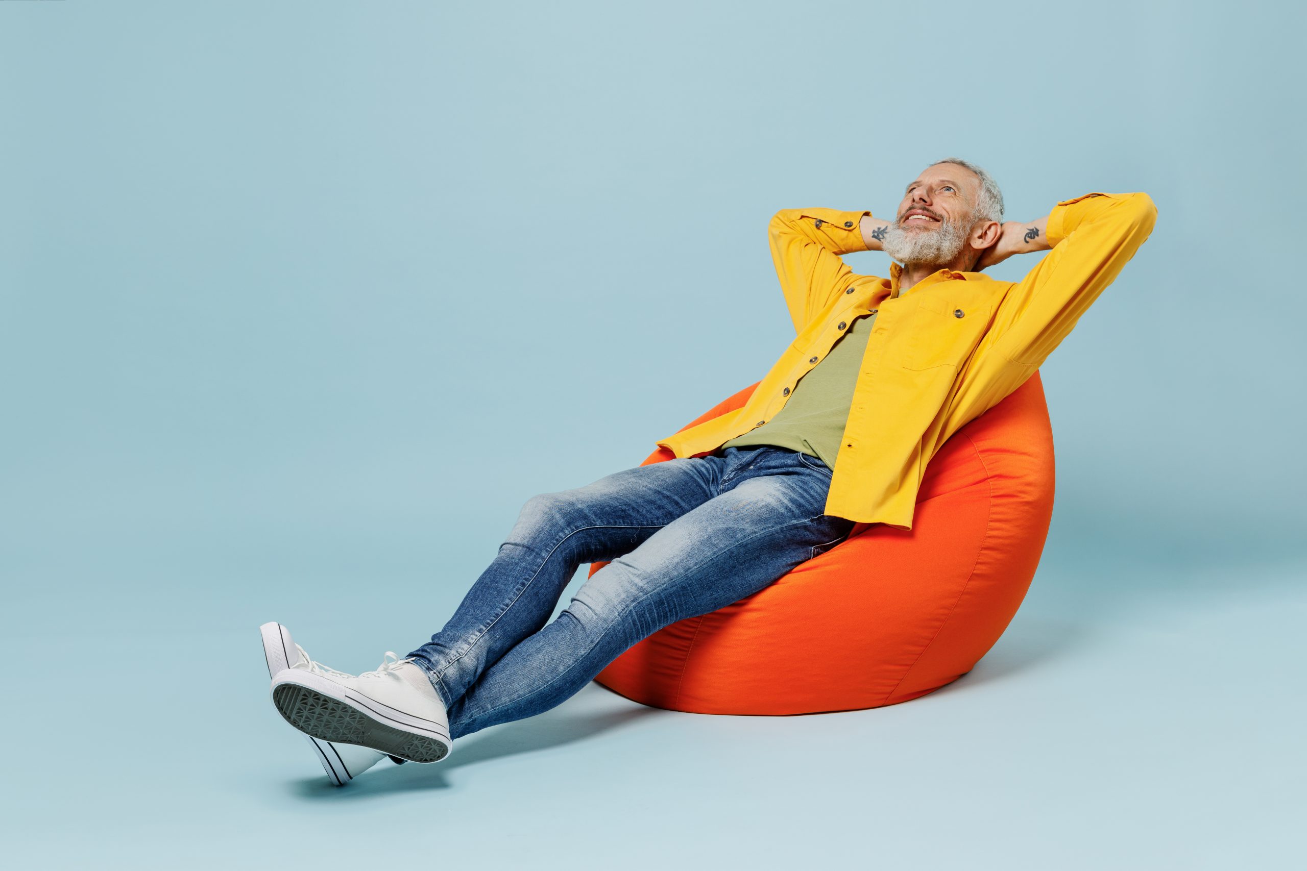 Full body sleeping elderly gray-haired mustache bearded man 50s in yellow shirt sit in bag chair hold hands behind neck isolated on plain pastel light blue background studio. People lifestyle concept