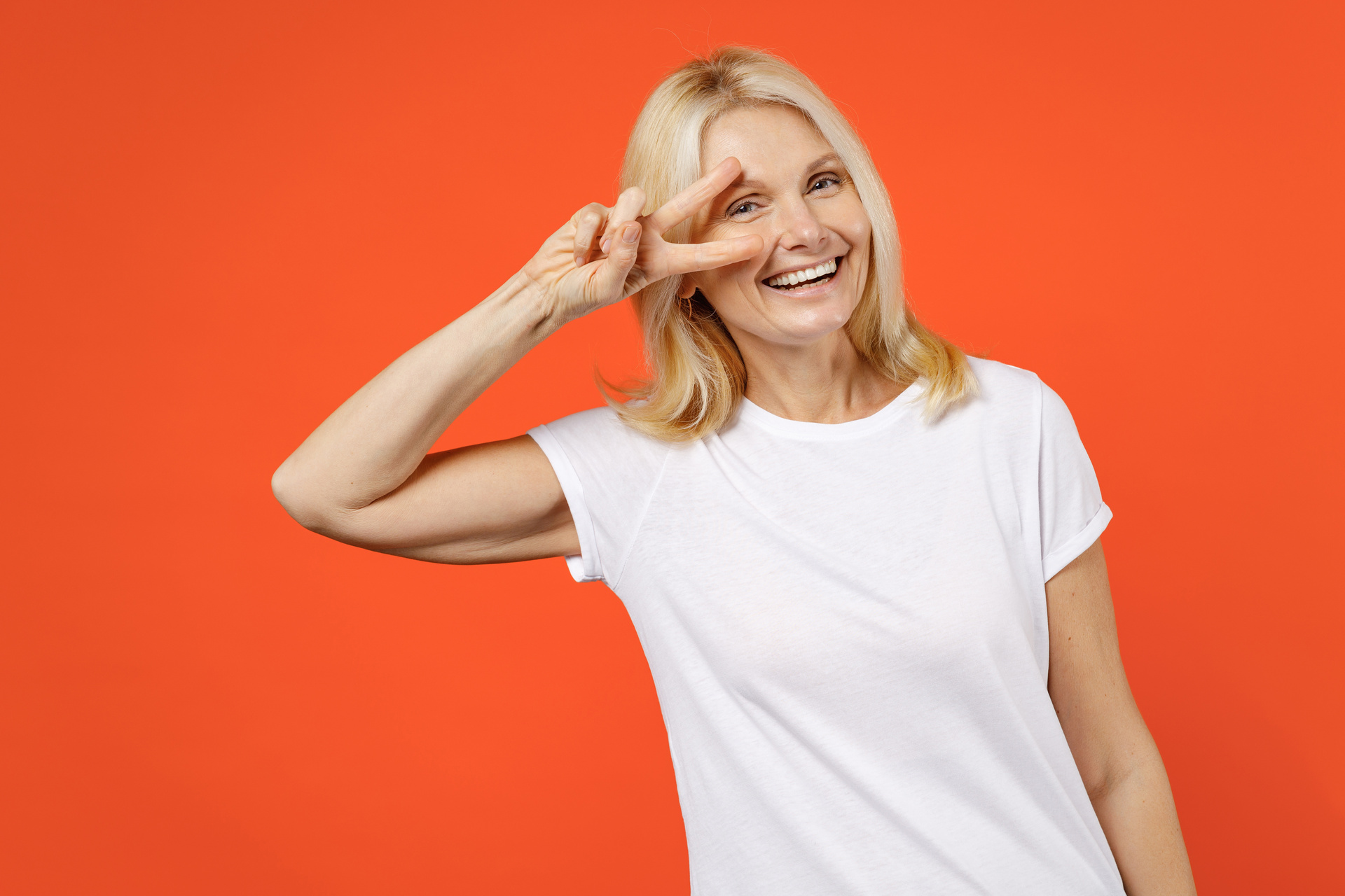Cheerful funny elderly gray-haired blonde woman lady 40s 50s years old in white casual t-shirt standing showing victory sign looking camera isolated on bright orange color background studio portrait