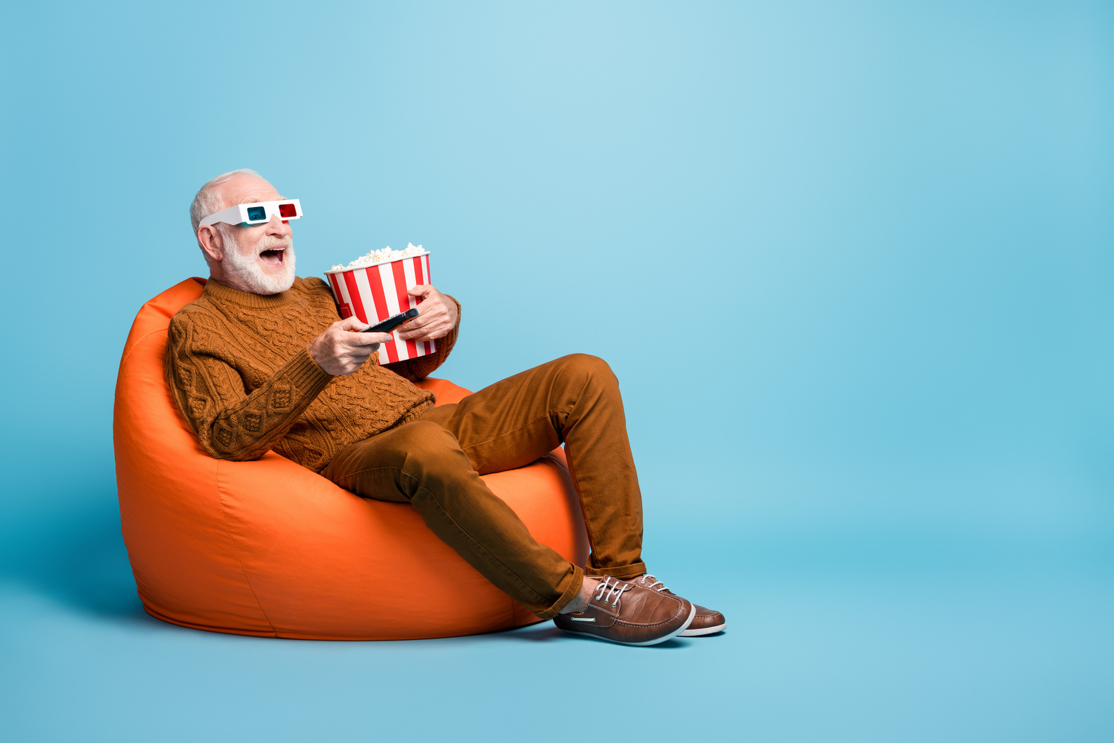 Portrait of his he nice attractive cheerful cheery bearded grey-haired man sitting in bag chair having fun eating corn watching tv comedy isolated over blue pastel color background